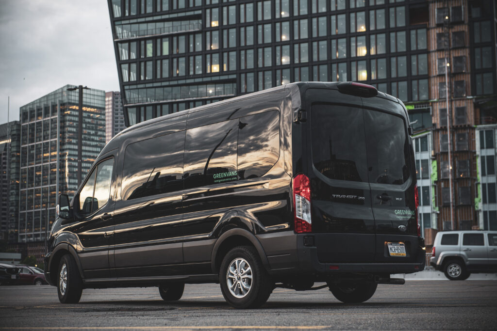 A black Ford Transit Passenger van rental with Greenvans parked in front of a high-rise office building.