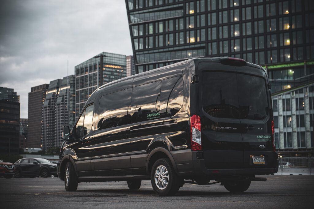 A black Ford Transit passenger van in front of a high-rise office building on a cloudy day