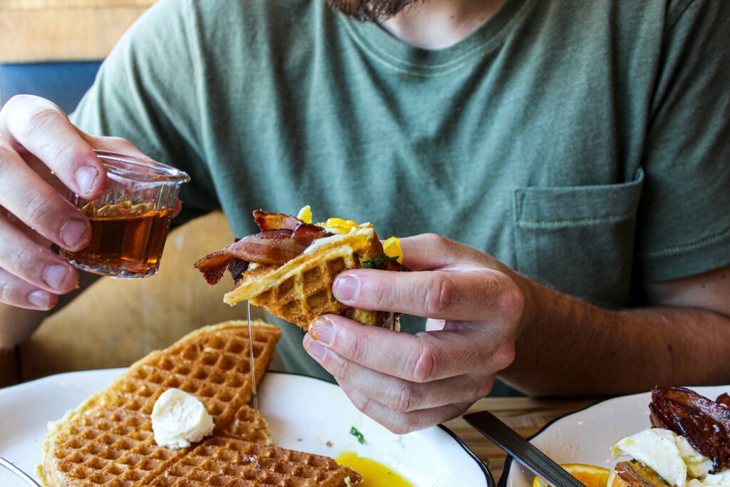 man eating breakfast at a diner of waffles, bacon and eggs, pouring syrup onto his food